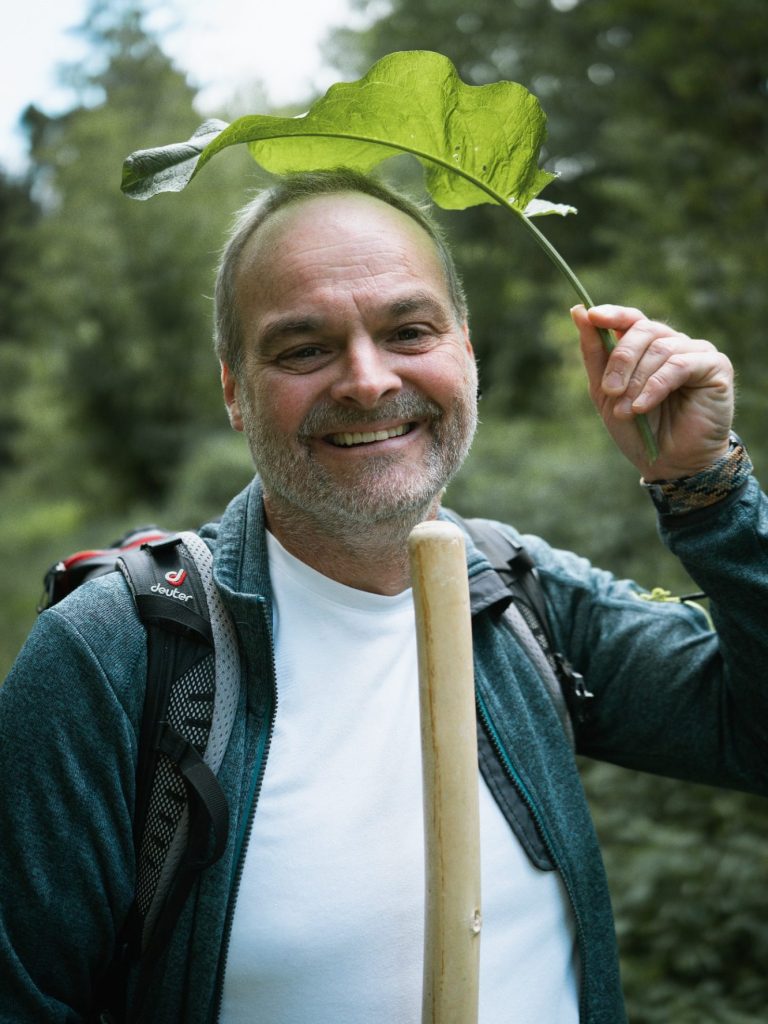 Naturcoach Dominic Zäh Mann mit einem Blatt als Hut, lächelt in einem Wald. Trägt einen Rucksack.